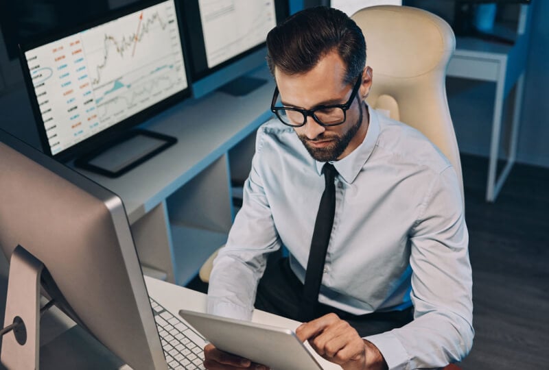 A man sitting at a desk with multiple monitors and interacting with a tablet device.